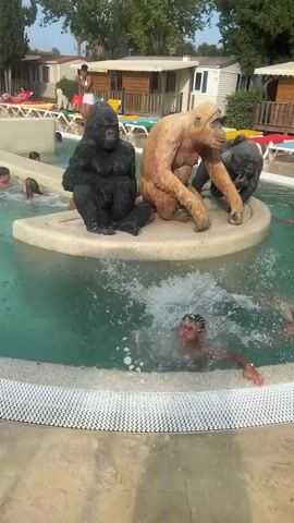 A boy in swim trunks splashes in a swimming pool. Three gorilla statues are on a circular platform in the pool, a black one on the left, a light brown one in the center, and a black one on the right. The boy is in the foreground, and the pool area extends into the background, with lounge chairs and mobile homes visible. The sun is shining.