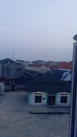 The video shows a slightly hazy, overcast sky, suggesting it might be early morning or late afternoon. The camera pans across a residential area with various buildings, predominantly houses and smaller structures. In the foreground, to the right, there's a glimpse of a metal railing and a portion of a building's facade. As the camera moves, it reveals a small building with a dark green roof and a light green trim around its base, possibly a mosque or a community center. Further back, there are other houses with tiled roofs in different colors, including red and grey. Some structures appear to be multi-story. Vegetation, including trees and shrubs, is visible interspersed among the buildings, and some areas show overgrown or unkempt grounds. The ground surface in the foreground and middle ground appears to be paved or made of concrete, with some areas showing signs of wear and tear. In the distance, a radio tower or antenna is visible against the sky. The overall impression is of a typical urban or suburban neighborhood.