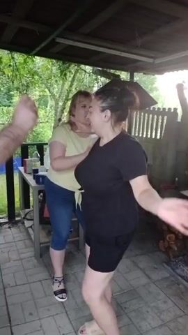 A short video shows a group of people at an outdoor gathering. In the foreground, a woman in a black t-shirt and shorts dances with her back to the camera, her arms raised. Next to her, a woman in a yellow t-shirt and blue jeans laughs. In the background, a man in a black t-shirt and shorts is visible on the left, also with his arms raised. Another man in a green t-shirt and grey shorts stands behind him. They appear to be on a covered porch or patio. A table with cups and bottles is visible to the left, and a pile of firewood is to the right. The overall impression is of a casual, festive atmosphere.