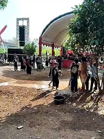 The video captures an outdoor gathering with a performance. In the foreground, a muddy area with puddles is visible. Several children, some dressed in black martial arts attire, are present. One child in the center is performing a martial arts move with a raised leg. Behind them, a group of adults and children watch the performance. A stage with a white canopy and red support poles is set up, where a performance appears to be taking place. Large speaker towers are positioned on the left side of the stage. The environment is outdoors, with trees and foliage visible in the background, suggesting a rural or semi-rural setting. The overall atmosphere seems festive or ceremonial.
