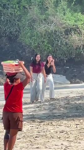 A busy beach scene unfolds under a bright sun. In the foreground, a person in a red shirt and brown shorts carries a pink basket on their head. Behind them, two young women in casual attire, one in a maroon top and the other in a black tank top, stand facing the water. The background reveals a rocky coastline with lush green trees and people swimming and playing in the ocean. Two empty lounge chairs and a large umbrella are positioned on the sand, suggesting a resort or relaxation area. The overall atmosphere is one of a vibrant and active beach environment.