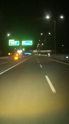 The video shows a first-person perspective from inside a moving vehicle on a multi-lane highway at night. The road ahead is illuminated by the vehicle's headlights and numerous streetlights overhead. On the left side of the highway, there are two green exit signs. The first sign, labeled "Keluar," indicates exits to "Seulimeum" and "Seulawah." The second sign directs to "Sigli" and "Lhokseumawe." Orange caution lights and reflective barriers are visible along the median separating the lanes. There are no other vehicles immediately in view, but faint lights from distant vehicles can be seen. The sky is dark and clear.