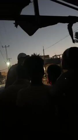 A group of people, silhouetted against a twilight sky, are gathered together. The camera angle suggests they are on a vehicle. In the foreground, the backs of several individuals are visible. One person in the mid-ground, wearing a cap, is facing away from the camera and appears to be speaking to someone. Beyond them, buildings and utility poles are discernible. The overall atmosphere is dim, with a streetlight illuminating the left side of the scene. The sky is a gradient of dark blue and purple, indicating either dawn or dusk.