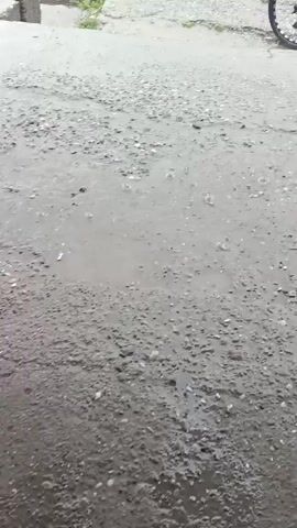 The video shows a cloudy, rainy day. Cars are parked on a wet asphalt surface. In the foreground, there is a broken concrete sidewalk with a drainage grate. In the background, there is a large building with "GLOBUS" written on it in green letters. Water is seen pouring from a pipe attached to a canopy structure, suggesting a malfunction or heavy rain. A person holding a plastic bag walks past the scene.