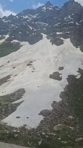 The video shows a mountain covered with snow, with patches of green grass and rocky terrain. The sky is blue with some white clouds. The mountain peaks are rocky and rugged, with some snow clinging to the higher elevations. The foreground is filled with more green vegetation and rocks. A song is playing in the background.