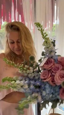 A woman with blonde hair stands in a well-lit room holding a large bouquet of flowers. She is wearing a pink skirt and a matching cropped sweater. She smiles as she looks at the bouquet. The room appears to be a flower shop or studio, with large windows, white curtains, and a large oval mirror in the background. There are plants and decorative elements scattered around the room. The woman is holding a mobile phone in her left hand. She looks as if she is posing for a picture or video, as she turns slightly to show off the bouquet.