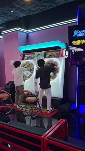 Two individuals are playing a large, dual-screen arcade game in a dimly lit entertainment venue. The game features bright, circular displays with moving graphics and lights. Both players are actively interacting with the game, one on the left in a white shirt and khaki pants, and the other on the right in a black shirt and white pants. They appear to be using their hands to touch or manipulate elements on the screens, suggesting a touch-based or motion-controlled game. Orange chairs are positioned in front of the arcade machine. The environment is characterized by neon lighting, with pink and blue accents. The floor is dark and reflective, and other arcade machines are visible in the background, along with seating areas. The overall atmosphere suggests a lively arcade setting.