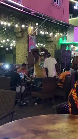 A man wearing a yellow basketball jersey with the number 23 on the back is standing and dancing in an outdoor seating area. He is facing away from the camera, with his hands raised above his head. Several other people are seated at tables, some are looking at the man. There are bottles of beer and red cups on the tables. The background is decorated with greenery and string lights.