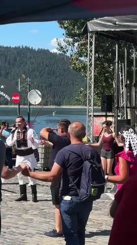 A group of people are dancing in a circle by a lake. A man in traditional clothing is singing into a microphone. There is a stage with speakers in the background. The weather is sunny.