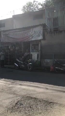 A street scene with a shop and a gas pump. The shop has a banner that reads "MADURA FAJAR". Several people are standing and sitting around the shop and the gas pump. A motorcycle is parked in front of the gas pump. The street is paved and cracked. The sun is shining and casting shadows.