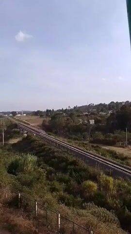 The video shows a view from an elevated position overlooking a rural landscape with a railway track running through it. The sky is pale blue with a few scattered clouds. The landscape features green vegetation, including bushes and trees, with some dry grassy areas.  The railway track extends into the distance, supported by a small bridge. Power lines run parallel to the track on the left side of the frame. In the distance, there are some buildings and what appears to be a small town or village. There is a metal fence in the foreground. The video appears to be a static shot with no camera movement.
