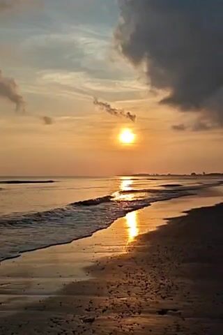 The video shows a beach scene during a sunset or sunrise. The sun is visible near the horizon, casting a bright reflection on the water. The waves are gently rolling onto the sandy beach. There are some clouds in the sky, and the overall color palette is warm and golden. The beach itself appears empty, with no people or objects immediately visible.