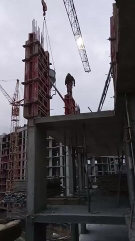 The video shows a construction site where a worker is standing on a structure made of red panels, guiding a crane that is lifting a bucket. Another crane is visible in the background, along with the unfinished framework of a large building. The sky is overcast, and the scene appears to be taken during the daytime.