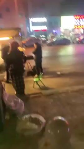 The video shows a nighttime view of a street in what appears to be an urban area. At the beginning, several individuals are standing around what seems to be a food cart with a light shining on it. The camera then pans and moves down the street, showing parked cars, businesses with neon signs, street lamps, and the fronts of buildings with windows and security cameras. The ground appears wet.