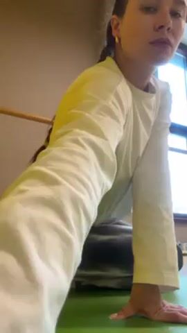 A young woman kneels on a green yoga mat near a barre in a room with a window in this video. She is fair-skinned with dark hair styled in a braid and is wearing a light yellow, long-sleeved shirt and gray tie-dye leggings. She looks to her left and then looks at the camera. The room appears to be a dance or exercise studio.