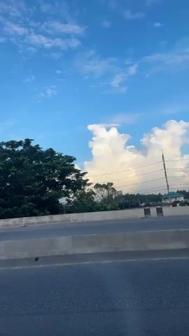 The video begins with a shot of a white semi-truck driving on a highway. The truck has the words "Don Adams" written on the side. A concrete barrier separates the highway lanes, and lush green trees line the roadside. The camera then pans upward to reveal a vivid blue sky dotted with puffy white clouds. Power lines are strung across the sky, weaving between the trees and clouds. The camera continues to pan, showcasing more of the cloudscape and a tree with white blossoms in the foreground. The overall environment suggests a bright, sunny day on a road trip.