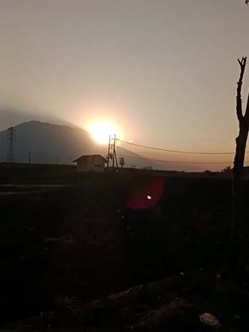 The video shows a sunset over a mountain. The sun is partially obscured by the mountain, casting rays of light across the sky. In the foreground, there is a field of dark earth, and in the middle ground, there is a small house with a power pole. There are also power lines visible in the sky. The sky is a pale orange and yellow, and the mountain is silhouetted against the light. The overall scene is one of natural beauty and tranquility.