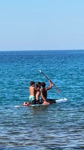 Three young males are on a paddleboard in the ocean. The male in the middle appears to be older and is sitting. The two younger males are kneeling, one on each side of the older male. The male on the left is leaning into the middle male, with his arm around his shoulder. The male on the right is holding a paddle. They are all facing away from the camera. The water is clear and blue, and the sky is clear and blue. The sun is shining.