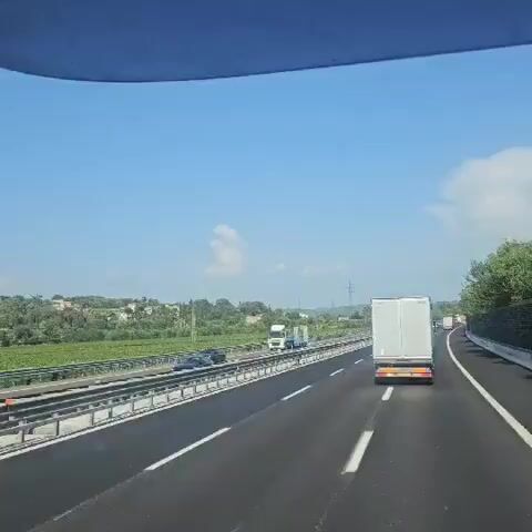 A dashcam view shows a highway with multiple lanes of traffic under a clear blue sky with scattered clouds. The camera is mounted inside a vehicle, likely a car, as evidenced by the windshield frame visible at the top. Several vehicles, including cars and semi-trucks, are traveling on the highway. On the left side of the road, there is a green field bordered by a metal barrier. Further in the distance, there are rolling hills covered with trees and some buildings. A large electricity pylon is visible on the right side of the road. The footage appears to be taken during daylight hours.