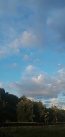 A handheld video shows a wide shot of a cloudy sky with some patches of blue visible. Trees and vegetation line the bottom of the frame, with a hint of a rural landscape in the distance. The camera is moving slowly upward, capturing the changing cloud formations.