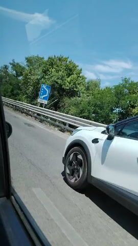The video shows a view from inside a moving vehicle, looking out the passenger window. A road with a metal guardrail on the right side is visible. Green trees and foliage line the road and extend into the distance under a clear blue sky with some scattered white clouds. Another car briefly appears, driving in the same direction, and then disappears from the frame. The video also captures a reflection of the vehicle's interior in the window.