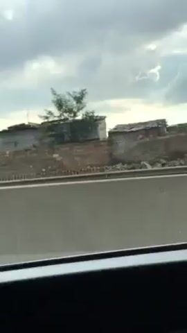 The video shows a moving car's-eye view of roadside scenery from a highway. The weather appears overcast. Adjacent to the highway, there is a settlement with simple structures, some built of brick or concrete blocks, others from corrugated metal. An advertising sign for a restaurant or food vendor is visible. Cars and trucks pass on the highway.