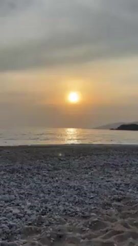 The video shows a beach at sunset. The sun is a bright, glowing orb in the sky, partially obscured by thin clouds, with its reflection shimmering on the water. The beach is covered in small, round pebbles in the foreground. The waves are gently lapping at the shore. A headland or distant landmass can be seen on the right side of the horizon.