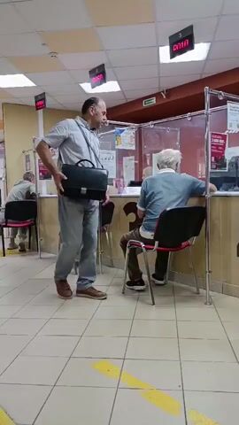 The video shows a queue of people in a public waiting area, possibly a government office or a bank. There are several counters with employees serving the customers. A man in a light blue shirt and jeans stands in the foreground holding a black briefcase. He appears to be waiting in line. In the background, other people are seated and standing, some interacting with the employees at the counters. The environment is well-lit with fluorescent lights, and the floor is tiled with yellow safety lines. The overall impression is one of a busy but orderly service center.