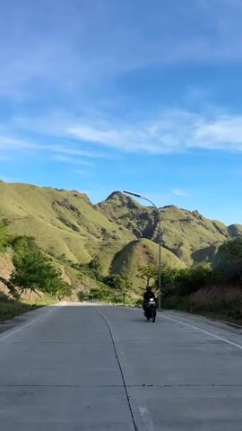 The video shows a motorcyclist riding down a concrete road towards the camera in a rural landscape. The road is straight, with a painted dividing line down the center and is flanked by green, grassy hills. A lamppost is visible on the right side of the road. The sky is blue with wispy clouds. The motorcyclist passes by, and the road is then empty.