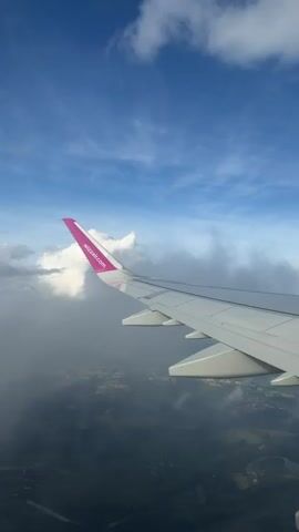 The video shows the wing of an airplane from a passenger's perspective. The wing is white with a pink tip that has the Wizz Air logo on it. Below the wing, the landscape is visible with patches of green and brown, suggesting a rural or suburban area with trees and fields. The sky is blue with white clouds, and sunlight is visible, indicating it is daytime. The airplane appears to be in flight.
