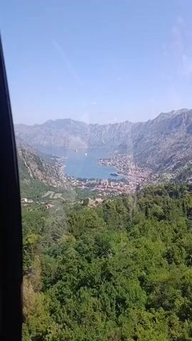 This media file captures a breathtaking panoramic view of the Bay of Kotor in Montenegro, likely from a high vantage point such as a cable car. The scene is dominated by the deep blue waters of the bay, dotted with several boats and a ferry. Nestled along the curved shoreline is the historic town of Kotor, with its distinctive red-tiled roofs and ancient architecture, appearing as a charming cluster of buildings.

The town is embraced by dramatic, rugged mountains that rise steeply on either side of the bay, creating a stunning fjord-like landscape. These mountains are covered in lush green vegetation, with dense forests of trees in the foreground and sparser greenery on the rocky slopes further back. The terrain suggests a mountainous coastal region.

The time of day appears to be daytime, indicated by the bright sunlight illuminating the scene and casting clear shadows, and the sky is a clear, vibrant blue, suggesting fair weather. The overall atmosphere conveyed is one of awe-inspiring natural beauty and the peaceful serenity of a picturesque coastal town. There are no visible people or specific objects that detract from the grandeur of the landscape. The view evokes a sense of tranquility and wonder, showcasing the harmonious coexistence of nature and human settlement.