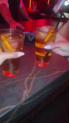 Two women are holding mixed drinks at a bar with a red light scheme. The women hold the drinks in front of themselves and move around, possibly toasting their drinks. The bar top is grey with a marbled effect. The music is upbeat and likely being played in a nightclub.