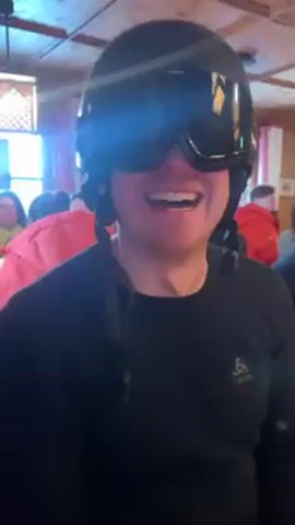 The video shows a person wearing a black ski helmet and goggles sitting inside what appears to be a restaurant or lodge. The person smiles and turns their head to the side, adjusting the helmet with their hand. The helmet has the brand name "RuRoC" on the side. The restaurant is crowded with people sitting at tables, some wearing ski jackets. The interior has a wooden ceiling and windows letting in light. There is also an ornate light fixture hanging from the ceiling. The camera pans slightly, showing more of the people and interior.