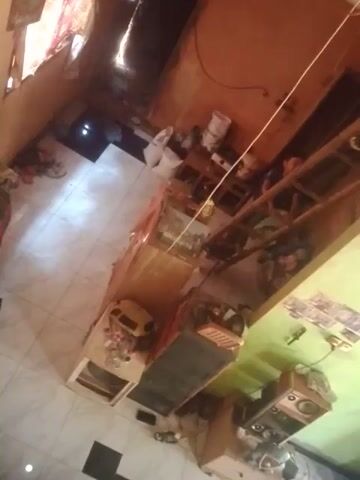 The video shows a room with light-colored tiled flooring. The room appears to be cluttered with various objects, including furniture, electronics, and what appear to be personal belongings. A long, thin, white string or cable runs diagonally across the scene from the upper left to the lower right. In the upper right corner, a beige wooden cabinet with speakers is visible. Below it, a lighter colored wooden cabinet is present. In the center of the room, a long wooden shelf unit with multiple compartments is filled with items. A glass-fronted cabinet or display case is situated near the center of the room. Another large, dark-colored electronic device is positioned to the left of the wooden shelf unit. Further towards the bottom of the frame, a white cabinet with drawers is visible. A yellow object, possibly a toy or appliance, is placed on a surface in the foreground. A person is partially visible in the upper left portion of the frame, lying down. The walls of the room are painted in different colors; one section appears light green, and another is an orange or brown hue. The lighting suggests it might be daytime or that artificial lights are on. The overall impression is of a lived-in, somewhat unorganized space.