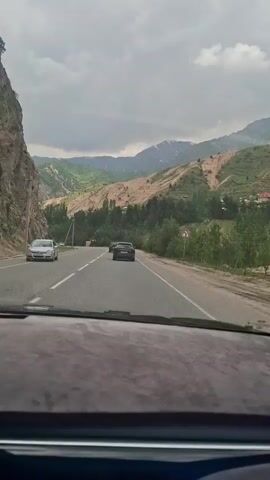 The video shows a car driving down a paved road in a mountainous region under a cloudy sky. There are three cars ahead of the camera. The road has two lanes, and there is a solid white line between them. On the left, there is a rocky cliff, and on the right, there are trees and green vegetation. In the distance, there are snow-capped mountains. A road sign is visible on the right. The dashboard of the car is visible in the foreground. There is music playing.