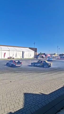 A group of cars are drifting in a square area outdoors. The area appears to be paved with cobblestones. In the background, there is a large white tent with a red banner hanging from it. There are also several orange traffic cones arranged in a circle on the ground, indicating a course or a designated area for the drifting. The sky is clear and blue, suggesting a sunny day. The cars are kicking up a lot of smoke as they drift.