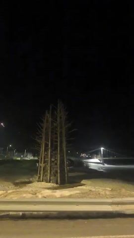 At night, a low-angle shot captures an illuminated bridge spanning a body of water. In the foreground, a sculptural arrangement of logs stands on a sandy or grassy area beside a road. Streetlights are visible along the road. The overall setting is dark, with the bridge lights providing the primary source of illumination.