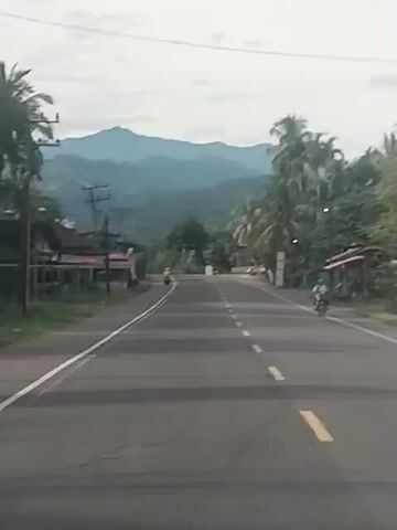The video shows a street scene with a road and buildings. A mountain range is visible in the background. A few people are on motorcycles. The road has a white line in the middle and a yellow line on the side. There are power lines and trees along the road. The sky is cloudy.
