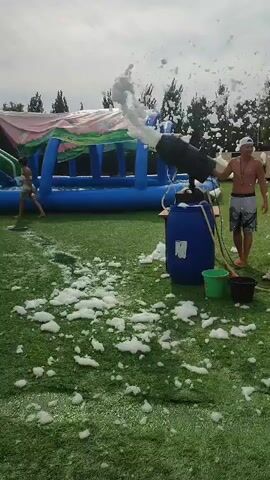 A man wearing a white cap and patterned shorts stands on a green artificial turf field. He is near a blue barrel with a green bucket and a black bucket next to it. A foam machine is mounted on top of the blue barrel and is actively spraying a large amount of white foam into the air. The foam is drifting across the field and onto the grass. In the background, there is a large inflatable structure that appears to be a water play area with blue and pink sections. Trees are visible in the far background, and the sky is overcast.