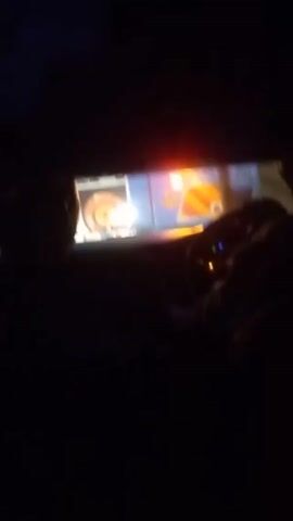 The video, taken from inside a vehicle, shows the rear of a blue and white bus with the license plate KDL 134F at night. The bus's tail lights and signal lights are illuminated. A person in a white shirt can be seen standing near the back of the bus. The driver of the vehicle is visible in the foreground, along with another person in the passenger seat. The overall environment appears dark, suggesting nighttime conditions.