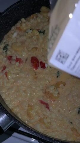 The camera focuses on a dark non-stick pan containing a mixture of small pasta, red and orange diced vegetables, and a light orange sauce. Someone is pouring a large amount of shredded or crumbled white cheese from a package into the pan. The cheese covers a significant portion of the existing mixture. A dark, slotted spatula is partially visible, embedded in the cheese-covered mixture. The pan is on a white stovetop.
