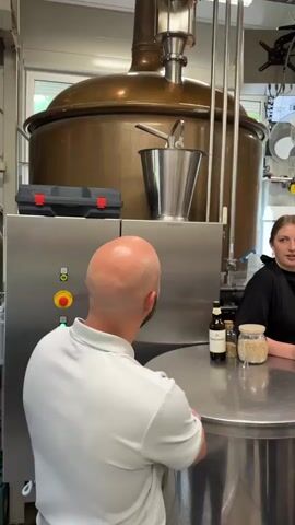 A medium shot captures a woman and a man in a modern brewery setting. The woman, on the right, wears a black top and has her hair tied back. She gestures with her hands as she speaks to the man, who is on the left with his back to the camera. He is bald and wears a white polo shirt, listening intently. A large copper brewing tank is visible in the background on the left, and a metal table with bottles, jars, and other brewing equipment is in the foreground. Pipes and machinery fill the rest of the background, along with a fan and a monitor.