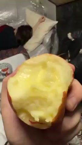 A hand holds a bitten apple with a light yellow interior. A person is lying down on a bed in the background, possibly sleeping. Behind the bed, there's a plastic covering or sheeting. A black trash bag is also visible in the background to the right. The person holding the apple takes another bite.