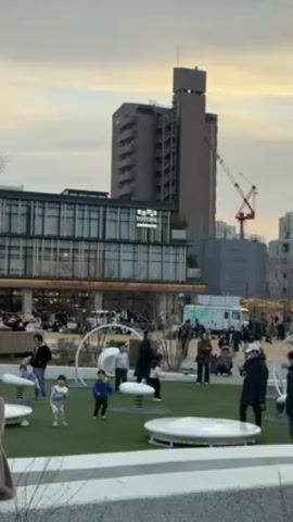 The video shows a public park in an urban setting. There is a building with the word "TOTOPA" visible on its facade. A tall, modern building is located behind the park, and a construction crane stands next to it. People are walking and children are playing in the park. The playground features modern, white play structures. The sky is overcast with an evening light.