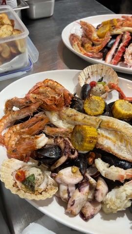 In a commercial kitchen setting, the video displays two plates of seafood. The primary plate, positioned closer to the camera, showcases a medley of grilled seafood, including shrimp, scallops, fish fillets, calamari, mussels, and grilled corn. The background plate features similar seafood elements. A person wearing black gloves and a wristwatch handles the primary plate, lifting it for display. The environment includes stainless steel surfaces and food storage containers.