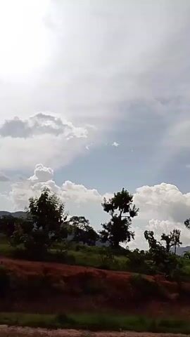 The video shows a landscape with a cloudy sky. In the foreground, there is a slope covered with red earth and green vegetation. Several trees are scattered across the landscape, with some appearing to be silhouetted against the bright sky. In the background, there are hills or mountains, also partially obscured by clouds. The sun is visible in the upper left corner of the frame, with bright rays filtering through the clouds. The overall scene depicts a natural, outdoor environment.