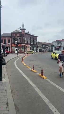 The camera is static, showing a wide view of a European city street on a cloudy day. 

A dedicated bike lane is evident in the foreground, distinguished by two parallel white lines and a series of evenly spaced yellow and red safety barriers. The asphalt road is slightly worn.

On the left, a building with a distinctive, ornate tower feature stands out, its dark brown and pink color scheme contrasting with the gray sky. Commercial establishments, including a pharmacy, line the ground floor. 

The street curves to the right, leading to a roundabout. A statue stands in the center of the roundabout, surrounded by a well-maintained grassy area bordered by red and white-painted curbs. Several cars are seen moving through the roundabout. 

Across the street, various commercial buildings and storefronts can be seen. A sign that reads "Amm" is prominently displayed on one of the buildings. Pedestrians are sparsely visible on the sidewalks. 

The overcast sky provides a diffused light, casting a muted tone over the scene.