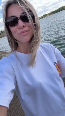 A woman with blonde hair and sunglasses takes a selfie outdoors near a body of water. She is wearing a white t-shirt. The water is rippling and there are trees and a cloudy sky in the background. The camera pans slightly throughout the video.