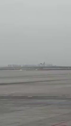The video shows an airplane landing on a gray tarmac, with city buildings visible on the horizon in the background. The sky is overcast.