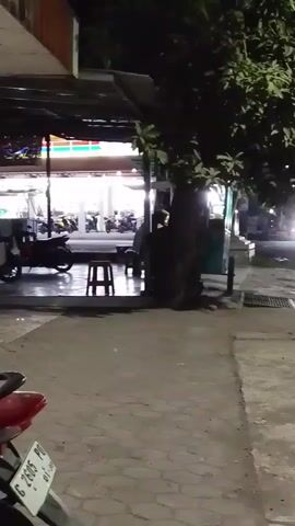 A nighttime view of a street scene, likely in an urban or suburban area. A motorcycle is parked in the foreground on the left, with a red tail light and a license plate visible. A sidewalk runs along the right side of the frame, with a tree growing by the street. Across the street, a brightly lit convenience store, possibly a 7-Eleven, is visible with several motorcycles parked outside. Traffic, including motorcycles and a car, moves along the street. The lighting suggests it is dark outside, with artificial lights from buildings and vehicles illuminating the scene.